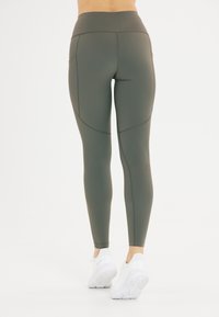 Opaque olijfgroene leggings met een hoge taille, voorzien van gebogen naden en perforatie-accenten nabij de kuit, gecombineerd met witte sportschoenen.