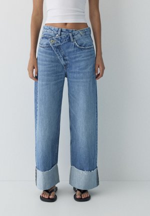 Kobieta nosząca jeansy z wysokim stanem, szerokimi nogawkami, dużymi mankietami i asymetrycznym paskiem z guzikami, połączone z czarnymi sandałami i białym krótkim topem.