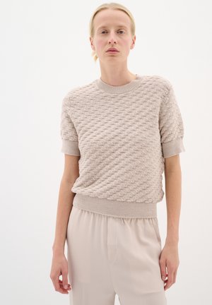 Pull en maille beige clair avec des manches courtes, présentant un col rond côtelé et un bas côtelé, associé à un pantalon doux et neutre.