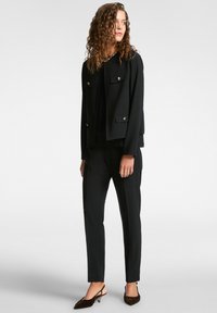 Chaqueta negra ajustada con dos bolsillos frontales y botones metálicos, combinada con pantalones slim-fit negros y zapatos oscuros con un tacón bajo.