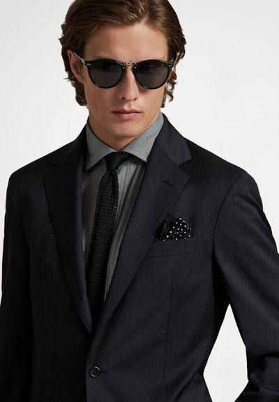 Mężczyzna w czarnym garniturze z wzorzystą poszetką, czarnym dzianinowym krawatem, szarą koszulą w kratę i okrągłymi czarnymi okularami przeciwsłonecznymi na białym tle.