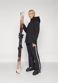Black ski jacket with a hood, black ski pants with white stripes, holding brown and black skis with bindings and a textured base.