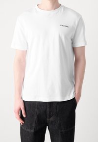 Bílý bavlněný tričko s kulatým výstřihem, krátkými rukávy a malým černým logem "Calvin Klein" na levé části hrudníku.