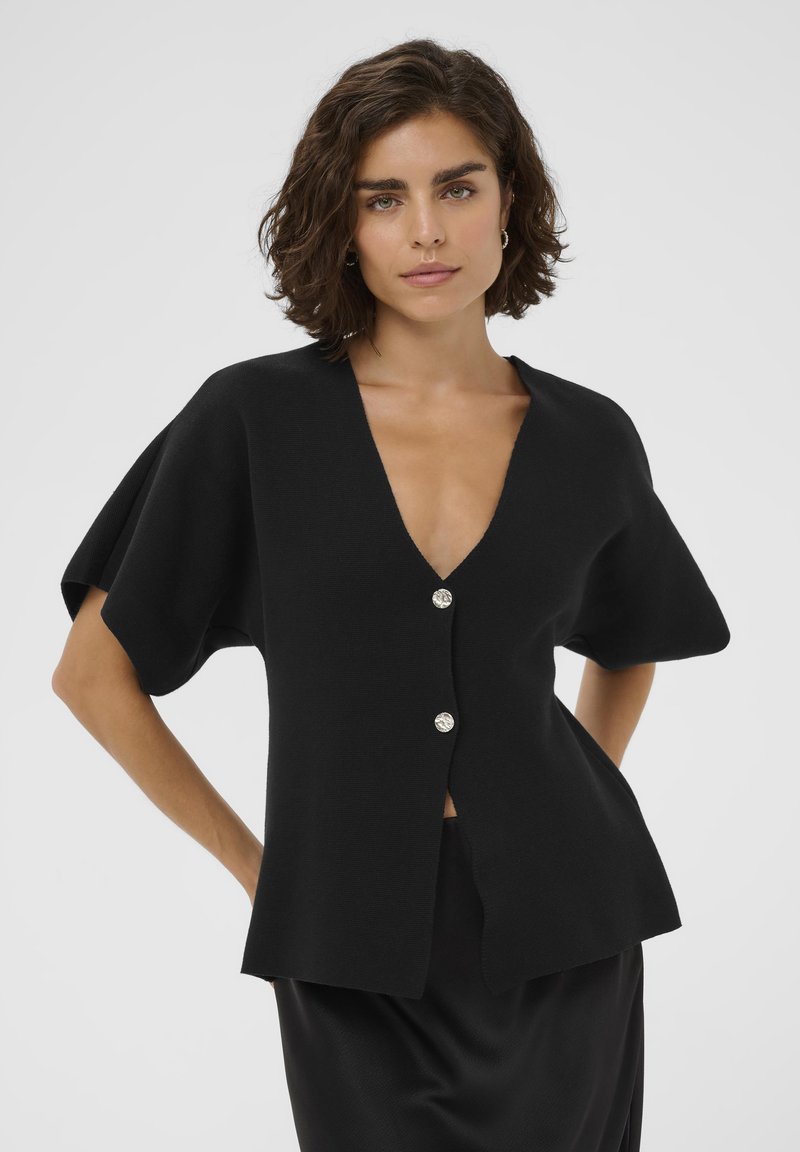 Gilet en tricot noir avec un décolleté en V profond, des manches courtes et deux boutons argentés à l'avant. Texture lisse, coupe structurée et design minimal.