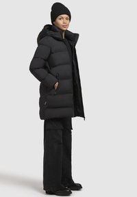 Jachetă puf cu glugă de culoare neagră, cu cusături orizontale, două buzunare laterale și o textură netedă. Asortată cu pantaloni cargo negri și cizme.