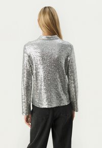 Sølvglitrende blazer med lange ærmer, en klassisk krave og lige snit. Kombineret med sorte bukser og set bagfra.