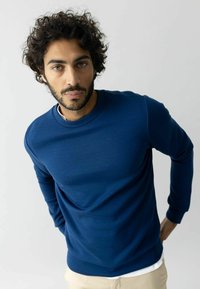 Tumši zils džemperis ar apaļu kaklu, ribinātām aprocēm un gludu tekstūru, uzvilkts virs baltas krekla, ar pieguļošu piegriezumu.