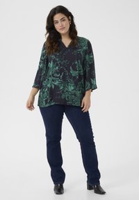 Blouse florale sombre en vert et marine, présentant un col en V et des manches 3/4, associée à un jean en denim foncé et des bottes noires à talons.
