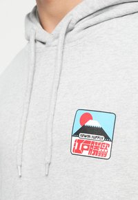 Sivá mikina s kapucňou, ktorá má na prednej strane nášivku s farebným dizajnom hory a slnka, s textom "EDWIN SUPPLY" v čiernej a červenej farbe.