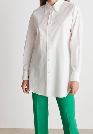 Henkilö, jolla on päällään pitkä valkoinen kauluksellinen nappipaita ja kirkkaanvihreät suorat housut, seisoo yksinkertaisen taustan edessä.