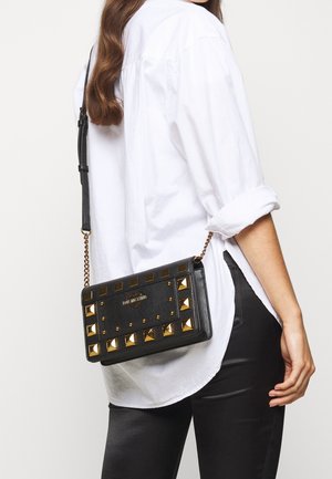 Sort læder crossover taske med gyldne nitter, rektangulær form, tekstureret overflade, kædestrop og præget logo på forsiden.