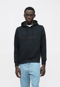 Vīrietis, kurš valkā melnu kapuci ar oranžu "JACK & JONES" tekstu un gaiši zilu džinsu, stāv uz vienkārša balta fona.