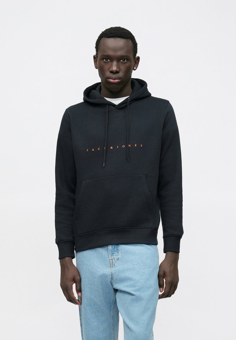 Vīrietis, kurš valkā melnu kapuci ar oranžu "JACK & JONES" tekstu un gaiši zilu džinsu, stāv uz vienkārša balta fona.