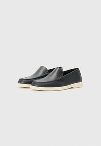 Mocassins noirs en cuir sans lacets avec bouts arrondis et semelles en caoutchouc blanches sur un fond gris clair uni.
