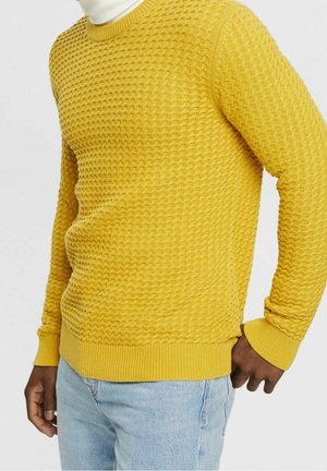 Man wearing textured yellow sweater over white turtleneck with light blue jeans, standing against plain background.