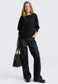 Zwarte, oversized trui met 3/4 mouwen, gecombineerd met hoge taille donkerdenim jeans en zwarte platform schoenen. Zwarte handtas inbegrepen.