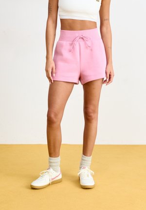 Vrouw draagt roze shorts met hoge taille, wit cropped tanktop, witte Nike-sneakers en lichtgekleurde sokken, staand op een gele vloer.