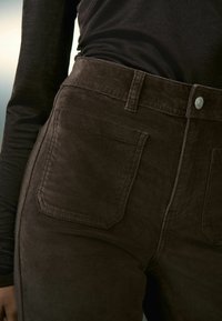 Pantalones de pana marrones con cintura alta, que cuentan con un cierre frontal de botón, dos bolsillos traseros y tejido texturizado para añadir detalle.