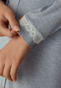 Ljusblå skjorta i en texturerad väv med intrikata vita spetsdetaljer vid ärmslutet, vilket framhäver fina detaljer och hantverk.