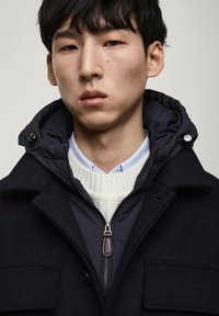 Svart ullkappa lagd över en marinblå puffjacka, med en vit stickad tröja, ljusblå skjorta med krage och en dragkedja framtill med detalj i metall.