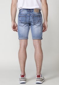 Pantalones cortos de mezclilla azul claro con puños enrollados, cinco bolsillos y una etiqueta de cuero en la parte posterior. Presenta detalles cosidos en los bolsillos.