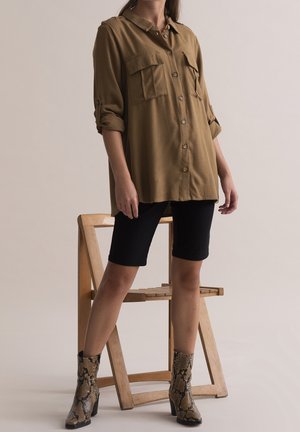 Personne portant une chemise marron à boutons, un short noir et des bottines à imprimé serpent, debout devant une chaise en bois.