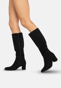 Botas altas de ante negras con tacón cuadrado, cremallera lateral y punta afilada. Textura suave con una silueta ajustada.