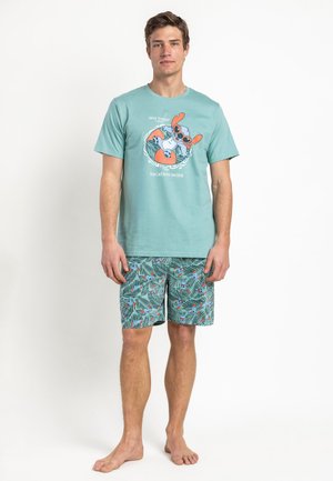 Joven de pie descalzo, lleva puestos unos pantalones cortos turquesa con estampado de hojas y pájaros y una camiseta turquesa con un perro de dibujos animados y el texto "Vacation Mode".