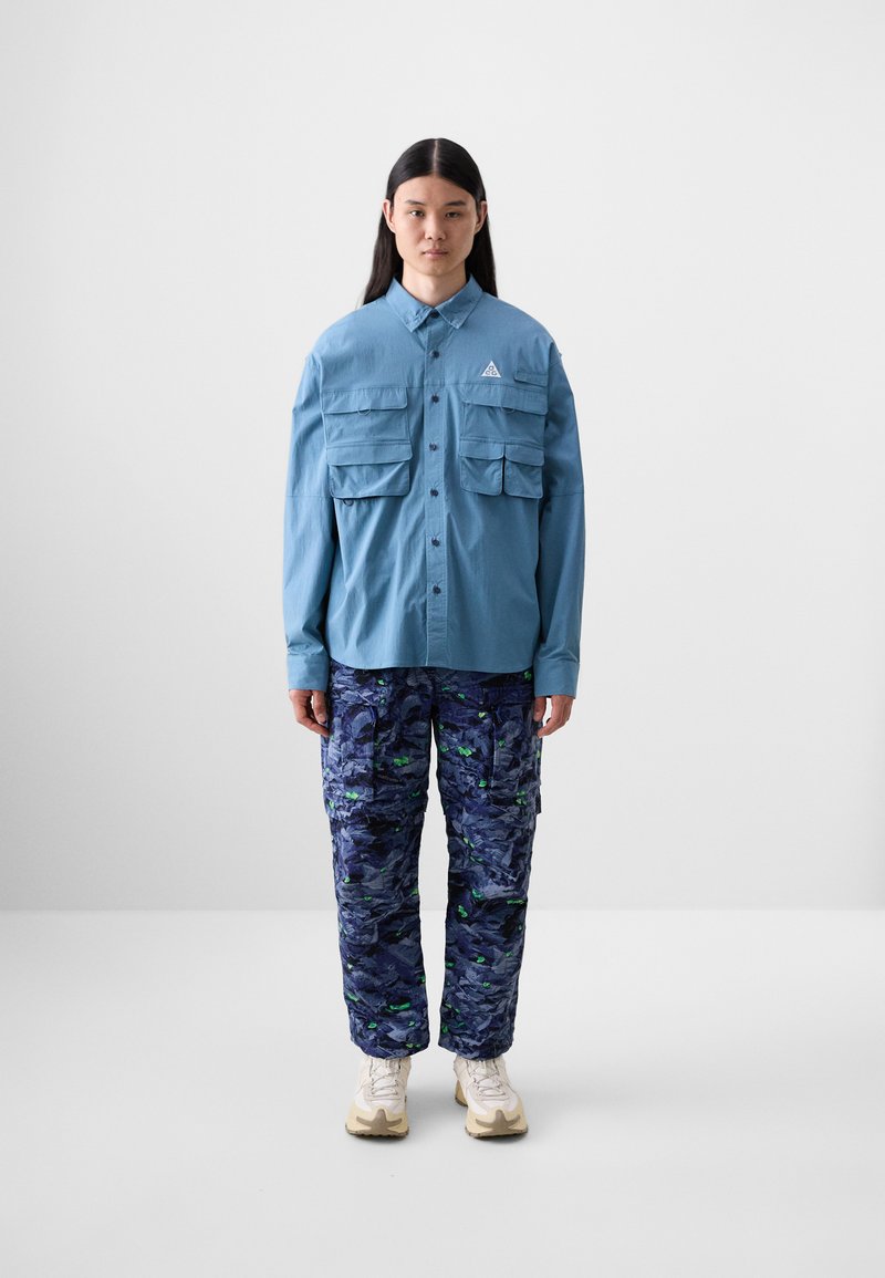 Lyseblå skjorte med knapp med to brystlommer; mørkeblå camo cargobukser med grønne detaljer; hvite joggesko.