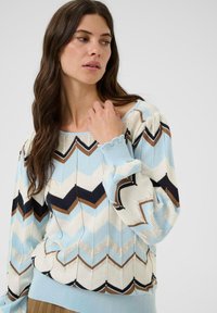 Femme avec de longs cheveux bruns portant un pull à motifs zigzag bleu clair, marron, blanc et noir, touchant le col, regardant sur le côté.