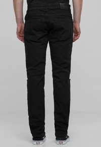 Jeans de mezclilla negra con un ajuste slim, que cuentan con dos bolsillos traseros y una textura suave. La cinturilla tiene una pequeña etiqueta; se llevan con zapatillas negras.