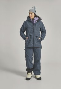 Dark blue waterproof jacket with hood, layered over a light purple mid-layer, paired with matching blue pants and cream snow boots.