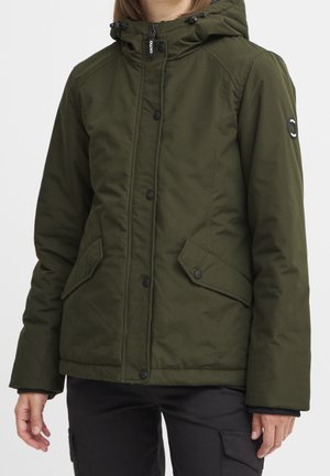 Femme portant une veste d'hiver à capuche vert olive avec des poches à rabat à boutons-pression et un pantalon cargo noir, debout devant un fond uni.
