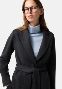 Blazer gris foncé avec une ceinture et deux poches avant, superposé sur un col roulé bleu clair. Le tissu semble doux avec une texture lisse.