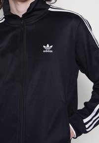 Svart zip-up jacka med hög krage, vit Adidas-logotyp på vänster bröst och tre vita ränder på varje axel. Slät tygtextur.