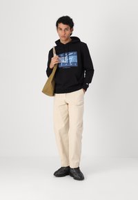 Sweat à capuche noir avec motif graphique, pantalon couleur crème et baskets noires. Le modèle tient un sac fourre-tout de couleur naturelle.