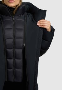 Melnā izolētā jaka ar augstu apkakli, kurai ir divkāršais rāvējslēdzējs, izšūta dizaina iekšējā slāņa un gluda ārējā tekstūra.