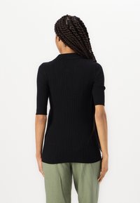 Black ribbed sweater with short sleeves and a collar, fitted shape, made of textured fabric, worn over green pants.