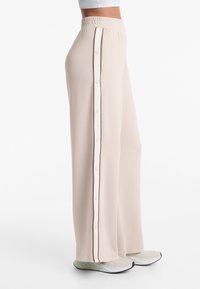 Helle beige weite Hosen mit samtiger Textur, die über einen seitlichen Knopfverschluss und kontrastierende dünne Paspelierung entlang der Nähte verfügt.