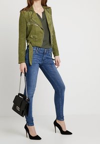 Olive green suede biker jacket with silver zippers, fitted blue jeans, and black high-heeled shoes. Black handbag with chain strap.