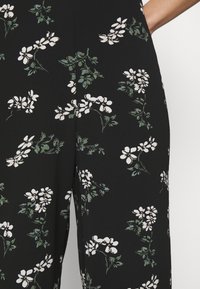 Tissu noir présentant un motif floral avec des fleurs blanches et des feuilles vertes. Le design est dense, créant un effet texturé.