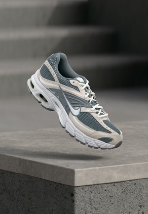 Grauer und weißer Nike-Laufschuh mit Mesh-Obermaterial und gedämpfter Sohle, schwebend über einem Betonsims vor einem unscharfen grauen Hintergrund.