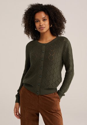 Frau mit lockigem Haar, die eine olivgrüne, geknöpfte Strickjacke und braune Velourshosen trägt und mit einer Hand in der Tasche vor einem einfarbigen Hintergrund steht.
