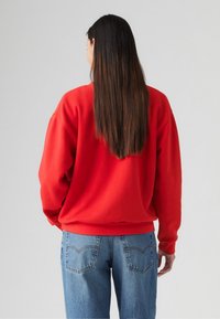 Felpa rossa con vestibilità rilassata, polsini e orlo a coste. Abbinata a jeans azzurri chiari con tasche posteriori e dettagli di cucitura.