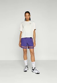 A nő egy egyszínű háttér előtt áll, fehér, oversized pólót, lila sportnadrágot narancssárga csíkokkal, fehér zoknit és fekete-fehér sportcipőt visel.