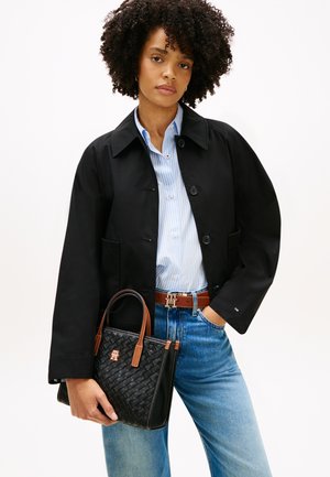 Kobieta o kręconych włosach, ubrana w czarną kurtkę, niebieską koszulę w paski, dżinsy, brązowy pasek, trzymająca plecioną czarną torebkę z brązowymi uchwytami.