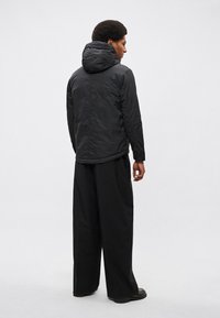 Μαύρο μπουφάν με κουκούλα και ματ φινίρισμα, με άνετη γραμμή, ορατά ραφές και συνδυασμένο με φαρδιά μαύρα παντελόνια. Απλός σχεδιασμός.