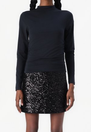 Femme portant un haut noir à manches longues ajusté et une mini-jupe noire à sequins, debout devant un fond blanc uni.