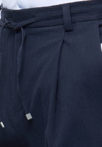 Σκούρα μπλε παντελόνια με υφή, ζώνη από ύφασμα και κορδόνια με μεταλλικές άκρες, σε συνδυασμό με ένα λευκό πουκάμισο που έχει μπει μέσα.