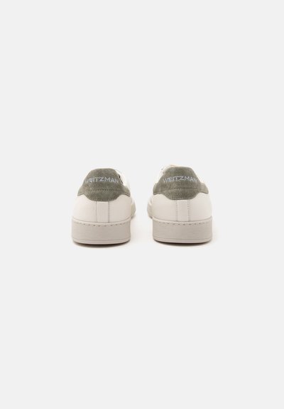 Paire de baskets blanches avec des languettes de talon en daim kaki brodées de "WEITZMAN", vues de dos sur un fond clair uni.